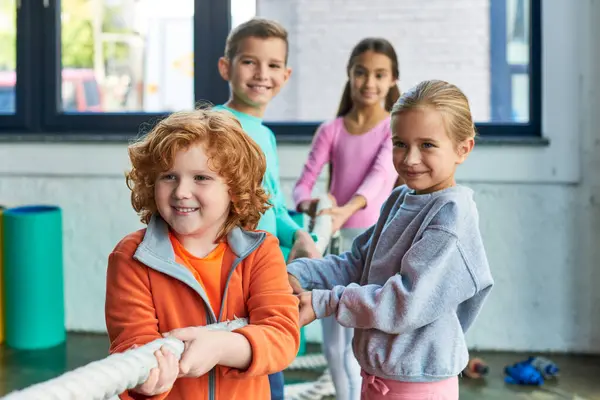 Cuatro niños pequeños y alegres en ropa deportiva jugando tirón de la guerra con cuerda de fitness, deporte infantil - foto de stock