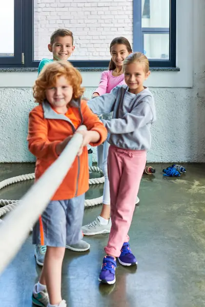 Tiro vertical de cuatro niños y niñas preadolescentes alegre jugando tirón de la guerra en el gimnasio, deporte infantil - foto de stock