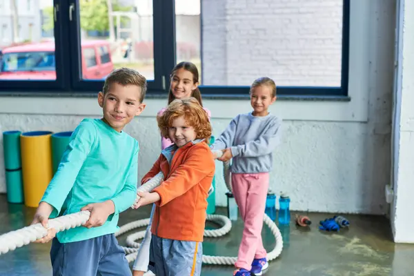 Alegres niños bonitos en ropa deportiva tirando de la cuerda de fitness en el gimnasio y sonriendo alegremente, deporte infantil - foto de stock