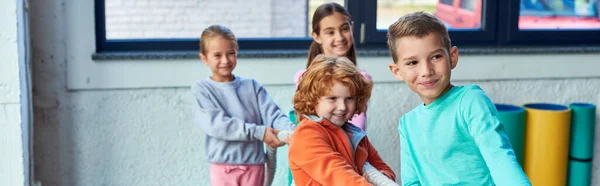 Niños felices en ropa deportiva tirando de la cuerda de fitness en el gimnasio y sonriendo alegremente, deporte infantil, pancarta - foto de stock