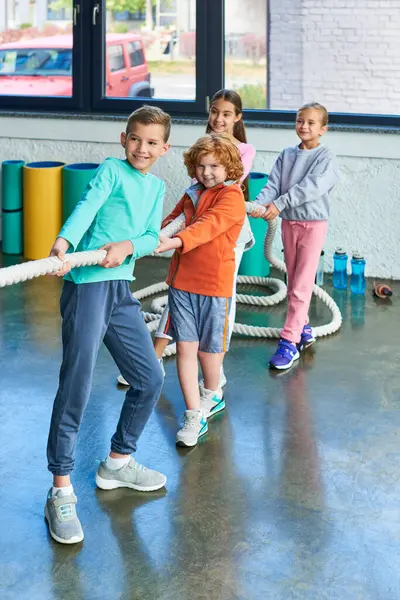 Tiro vertical de pequeños niños alegres tirando de la cuerda de fitness y sonriendo sinceramente, deporte infantil - foto de stock