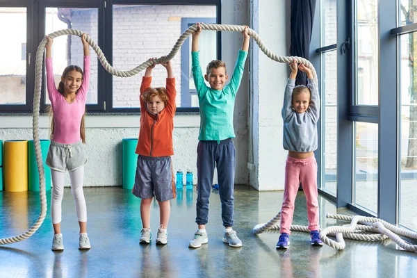 Niños alegres en ropa deportiva elevar la cuerda de fitness por encima de las cabezas y sonreír a la cámara, deporte infantil - foto de stock