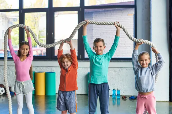 Cuatro pequeños niños lindos en ropa deportiva elevar la cuerda de fitness por encima de sus cabezas, deporte infantil - foto de stock