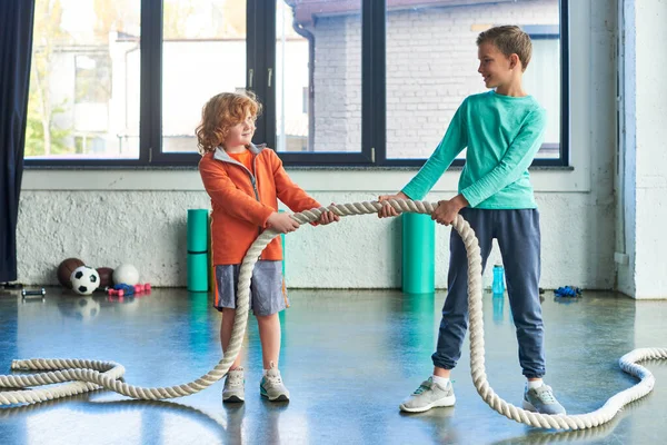 Red haired and brunette boys in sportswear pulling fitness rope and looking at each other, sport — Stock Photo