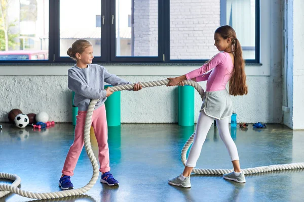 Веселі маленькі дівчата в спортивному одязі тягнуть фітнес-канат і радісно посміхаються один одному, спорт — стокове фото