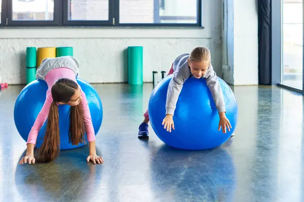 Dos niñas alegres en ropa deportiva acostado en pelotas de fitness en el gimnasio estirando las manos al suelo, deporte infantil - foto de stock
