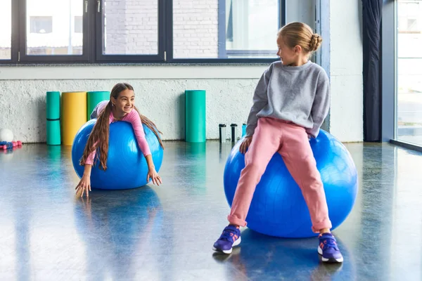 Duas meninas pequenas alegres exercitando-se em bolas de fitness e sorrindo um para o outro, esporte infantil — Fotografia de Stock
