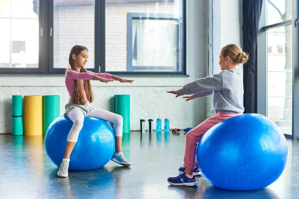 Zwei hübsche kleine Mädchen, die auf Fitnessbällen sitzen und die Arme nach vorne strecken, Sport treiben — Stockfoto