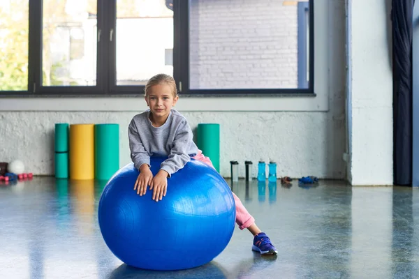 Hübsches kleines Mädchen in Sportbekleidung auf Fitnessball liegend und leicht lächelnd in die Kamera, Kindersport — Stockfoto