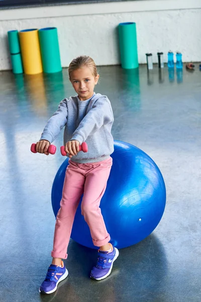 Bonita niña en ropa deportiva sentada en la pelota de fitness y haciendo ejercicio con pesas, deporte infantil - foto de stock