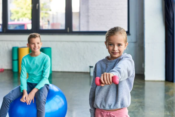 Vorpubertierendes blondes Mädchen trainiert mit Kurzhantel und kleinem Jungen auf Fitnessball im Hintergrund — Stockfoto