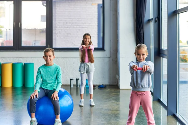 Niedlicher kleiner Junge sitzt auf Fitnessball mit zwei hübschen Mädchen, die mit Hanteln üben, Kindersport — Stockfoto