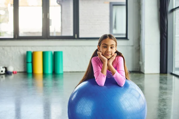 Alegre niña con el pelo largo en ropa deportiva rosa posando con pelota de fitness, las manos bajo la barbilla - foto de stock