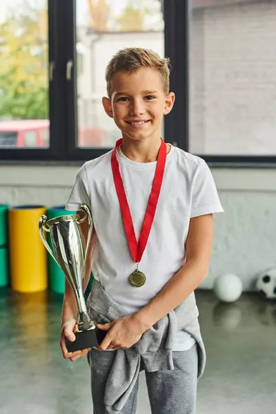 Tiro vertical de niño lindo con medalla de oro y trofeo sonriendo a la cámara, deporte infantil - foto de stock
