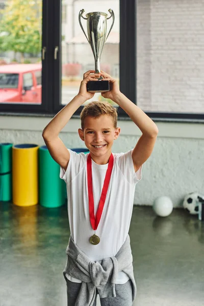 Vertical shot of cheerful little boy with golden medal raising trophy above his head, child sport — Stock Photo