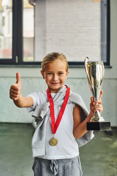 Senkrechte Aufnahme eines blonden Mädchens mit Trophäe und Goldmedaille, Daumen hoch in die Kamera, Kindersport — Stockfoto