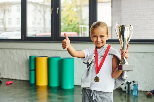 Niña rubia posando con trofeo y medalla de oro y mostrando el pulgar hacia arriba en la cámara, deporte infantil - foto de stock