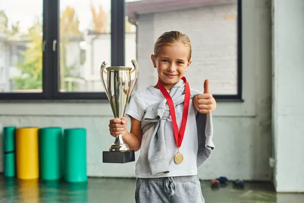 Alegre niña preadolescente con medalla de oro que muestra el pulgar hacia arriba y la celebración de trofeo, deporte infantil - foto de stock