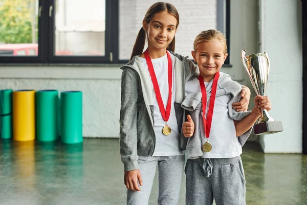 Dos chicas preadolescentes lindas con medallas sosteniendo trofeo, sonriendo a la cámara, pulgar hacia arriba, deporte infantil - foto de stock