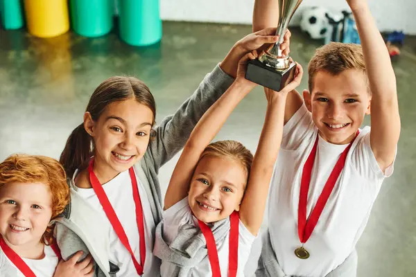 Cuatro alegres niños preadolescentes lindos en ropa deportiva triunfando con un enorme trofeo en las manos - foto de stock