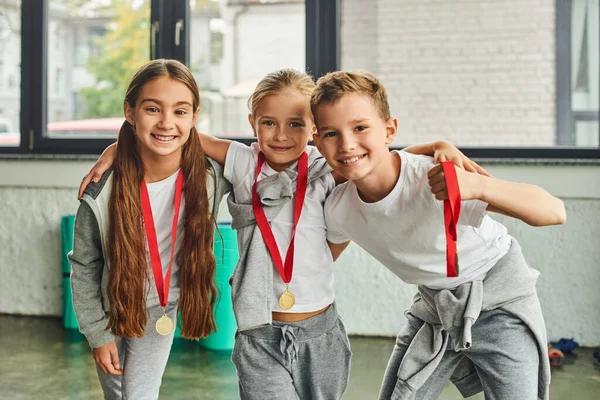 Three little friends in sportswear with golden medals hugging and smiling at camera, child sport — Stock Photo