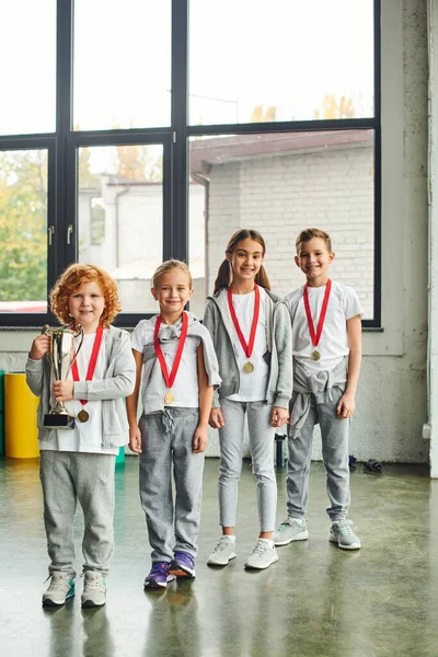 Вертикальный снимок маленьких мальчиков и девочек, позирующих с золотыми медалями и трофеями, смотрящих в камеру — стоковое фото
