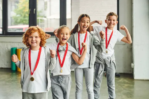 Cuatro niños alegres en ropa deportiva con medallas animando y sonriendo felizmente a la cámara, deporte infantil - foto de stock