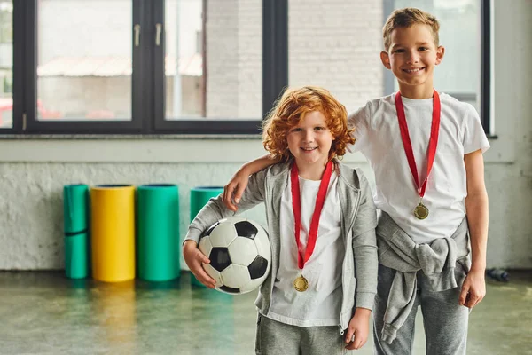 Two cheerful boys in sportswear with golden medals holding soccer ball and smiling at camera, sport — Stock Photo