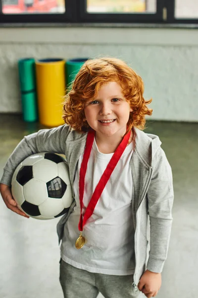 Tiro vertical de menino ruivo bonito com medalha de ouro segurando bola de futebol e sorrindo alegremente — Fotografia de Stock