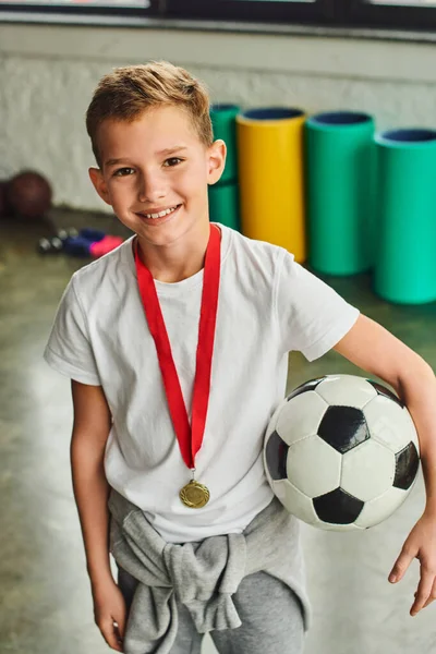 Vertical shot of little boy with golden medal and soccer ball with fitness mat on backdrop, sport — Stock Photo