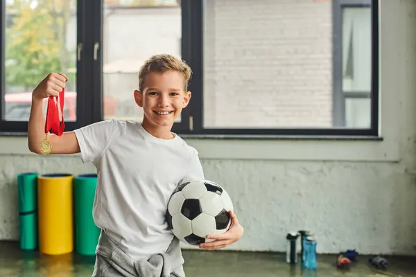 Niedlich fröhlicher vorpubertärer Junge in Sportkleidung mit Goldmedaille und Fußball, Kindersport — Stockfoto