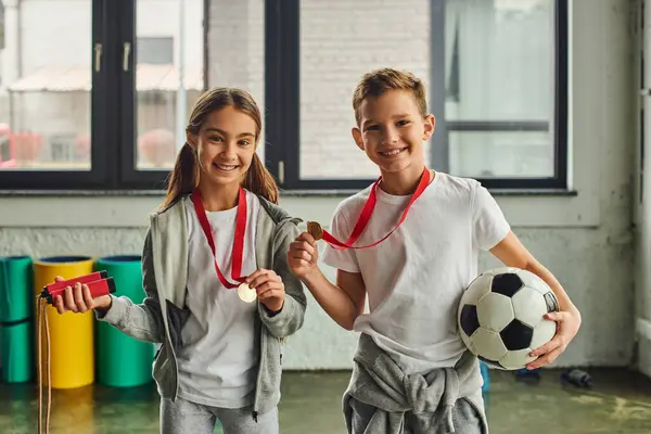 Little girl with medal holding jump rope and cute boy posing with soccer ball, smiling at camera — Stock Photo