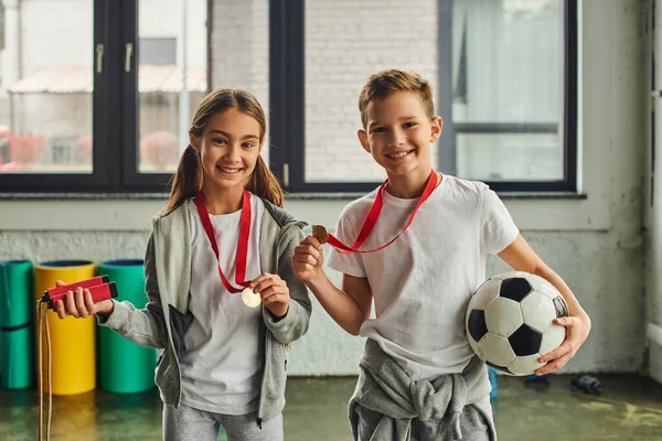 Menina com medalha segurando corda de salto e menino bonito posando com bola de futebol, sorrindo para a câmera — Fotografia de Stock