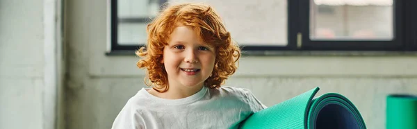 Carino piccolo ragazzo dai capelli rossi in posa con karemat in mano e sorridente alla macchina fotografica, sport bambino, banner — Foto stock