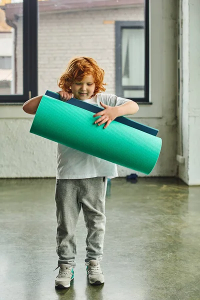 Tiro vertical de menino bonito com cabelo vermelho em sportswear desdobrando seu karemat, esporte infantil — Fotografia de Stock