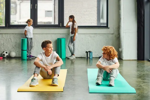 Dois meninos alegres sorrindo um para o outro em tapetes de fitness com garotas bonitos de pé no fundo — Fotografia de Stock
