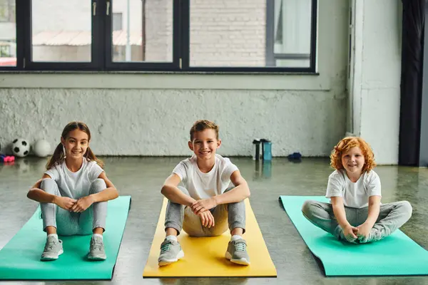 Drei fröhliche vorpubertäre Jungen und Mädchen, die auf Fitnessmatten sitzen und in die Kamera lächeln, Sport treiben — Stockfoto
