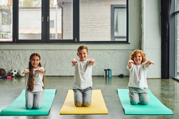Pequeños niños alegres sentados en colchonetas de fitness y brazos extendidos, sonriendo a la cámara, deporte infantil — Stock Photo