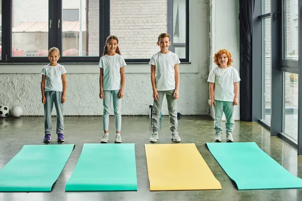 Jolly preadolescent boys and girls in sportswear posing next to fitness mats in gym, child sport — Stock Photo