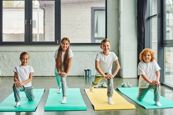 Allegre bambine e ragazzi che allungano le gambe sui tappetini fitness e sorridono gioiosamente, sport infantile — Foto stock