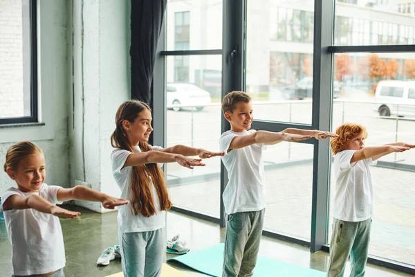 Crianças pequenas alegres em sportswear esticando os braços no ginásio com fundo de janela, esporte infantil — Fotografia de Stock