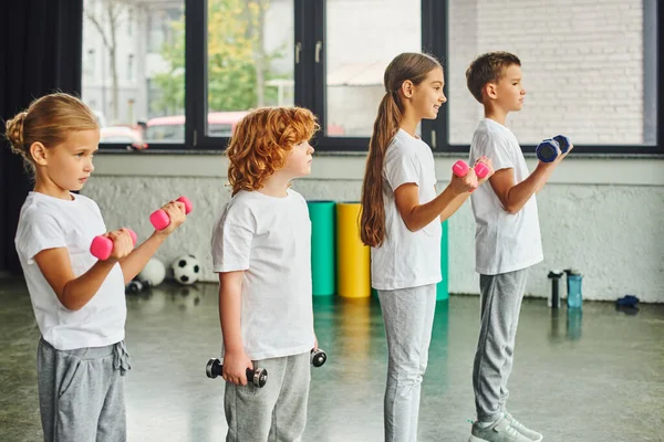 Niños pequeños posando en perfil sosteniendo pesas con colchonetas y pelotas de fitness sobre el telón de fondo, deporte - foto de stock