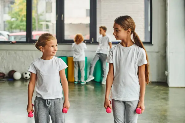 Zwei kleine Mädchen mit Hanteln, die einander anschauen, mit verschwommenen Jungen auf dem Hintergrund, Kindersport — Stockfoto