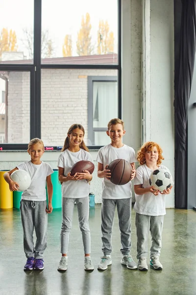 Tiro vertical de cuatro niños preadolescentes con diferentes tipos de bolas, sonriendo a la cámara, deporte - foto de stock