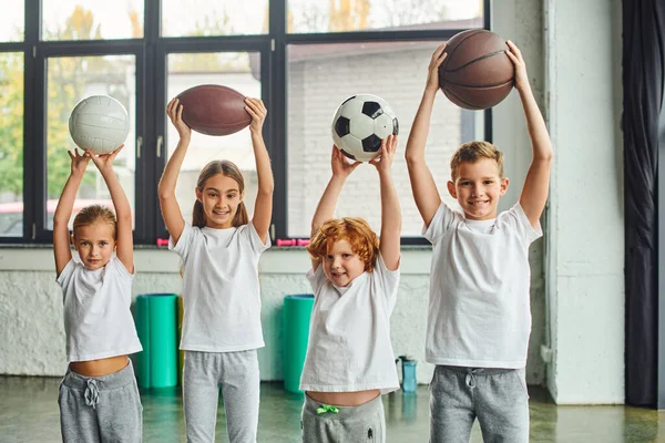 Meninos e meninas alegres posando com diferentes tipos de bolas acima de suas cabeças, esporte infantil — Fotografia de Stock