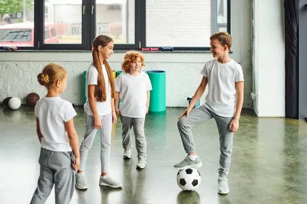 Meninas bonitos e vermelho peludo menino sorrindo para outro menino com perna na bola de futebol, esporte infantil — Fotografia de Stock