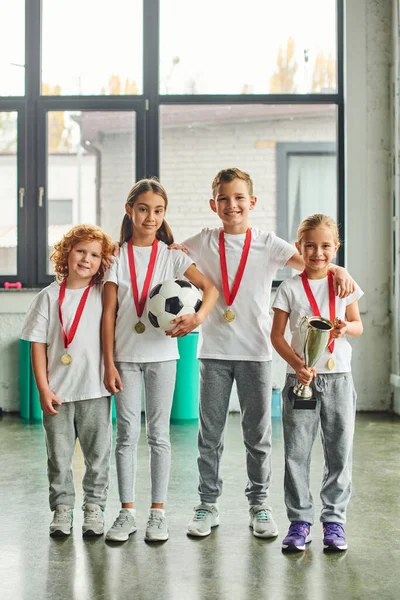 Vertical shot of preadolescent cute children with golden medals posing with soccer ball and trophy — Stock Photo