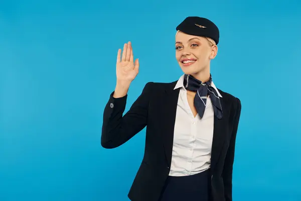 Jeune hôtesse accueillante en uniforme professionnel élégant agitant la main sur fond de studio bleu — Photo de stock