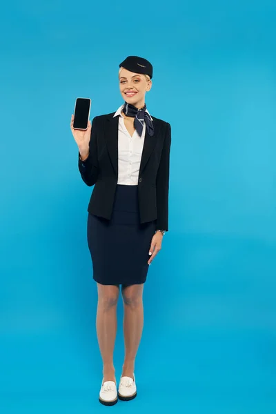 Azafata sonriente en elegante uniforme que sostiene el teléfono inteligente con pantalla en blanco sobre fondo azul - foto de stock