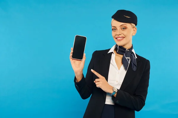 Elegante azafata en elegante uniforme apuntando al teléfono inteligente con pantalla en blanco en el telón de fondo cian - foto de stock